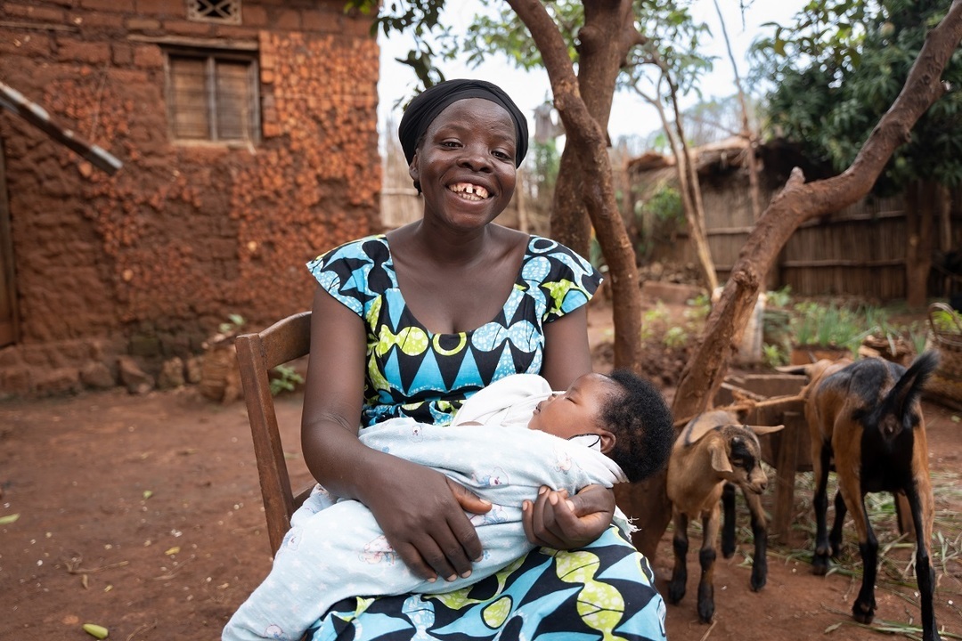 A woman sits on a chair with her baby in her arm in front of her house. A few goats in the background (Source: Christian Nusch)